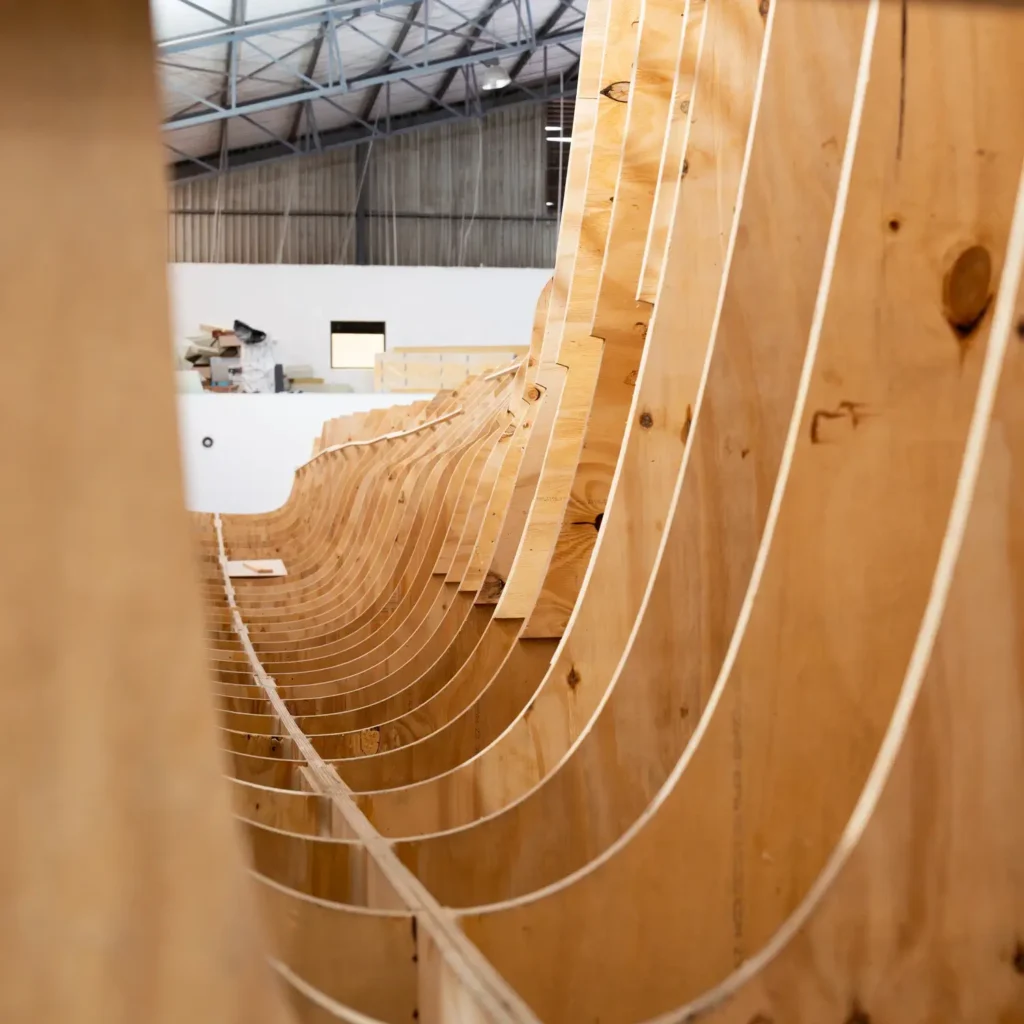 Wooden ribs of a boat hull under construction in a workshop, with visible curved frames and a partially built structure in the background.