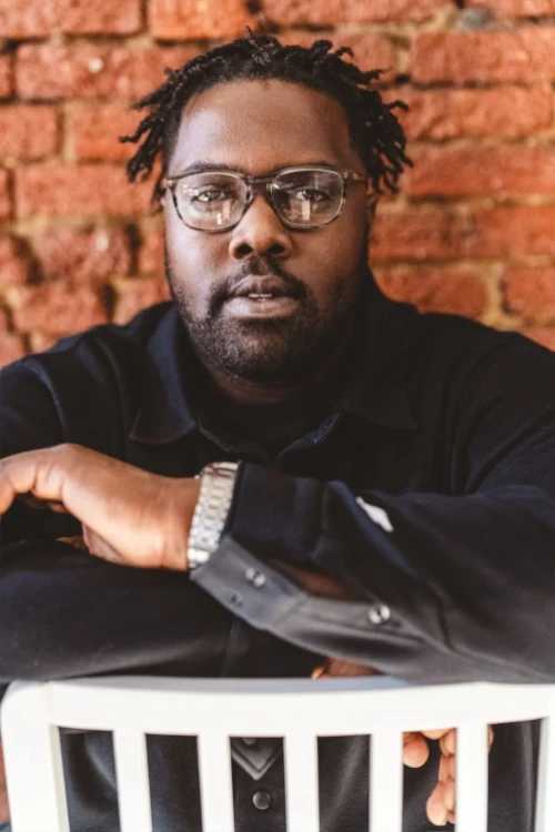 A man with glasses and short dreadlocks sits backward on a white chair, resting his arms on the chair back, in front of a red brick wall.