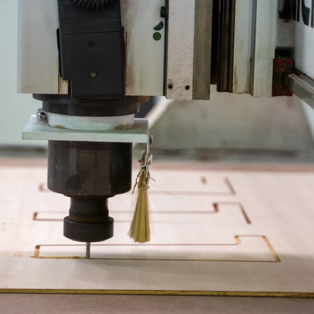A CNC machine router cuts precise shapes into a sheet of wood, with a small cleaning brush attached near the cutting tool.