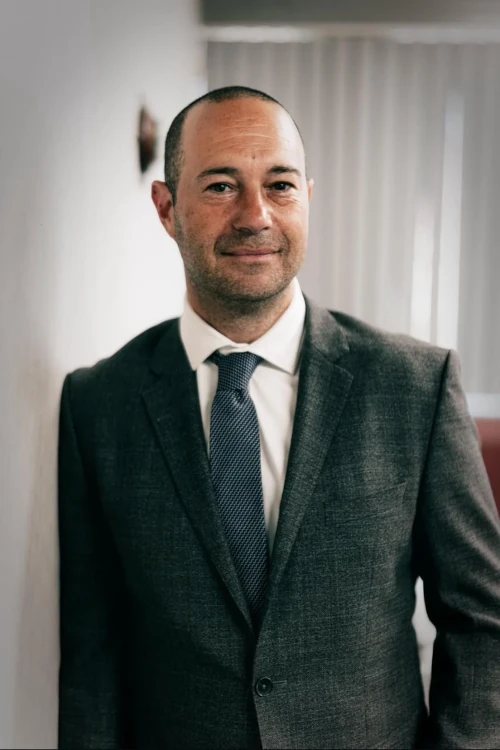 A man in a gray suit and blue tie stands indoors, leaning slightly against a white wall, with vertical blinds in the background.