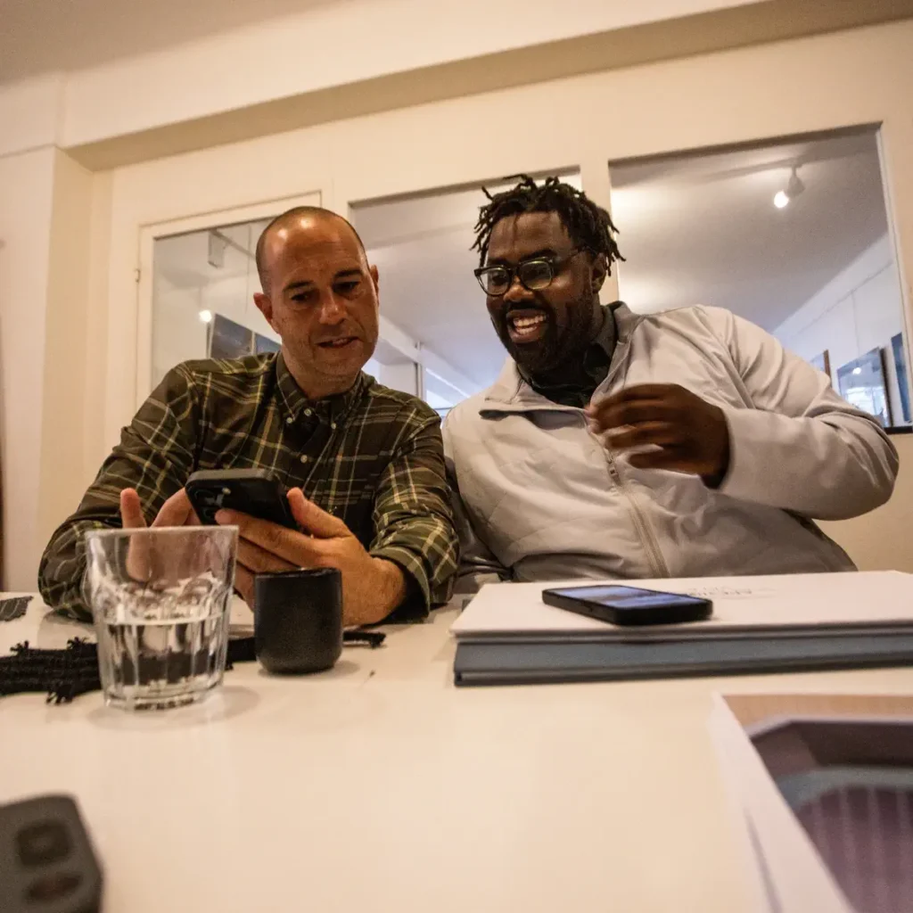 Two men sitting at a table look at a smartphone and smile. There are glasses of water, a notebook, and a phone on the table in front of them.